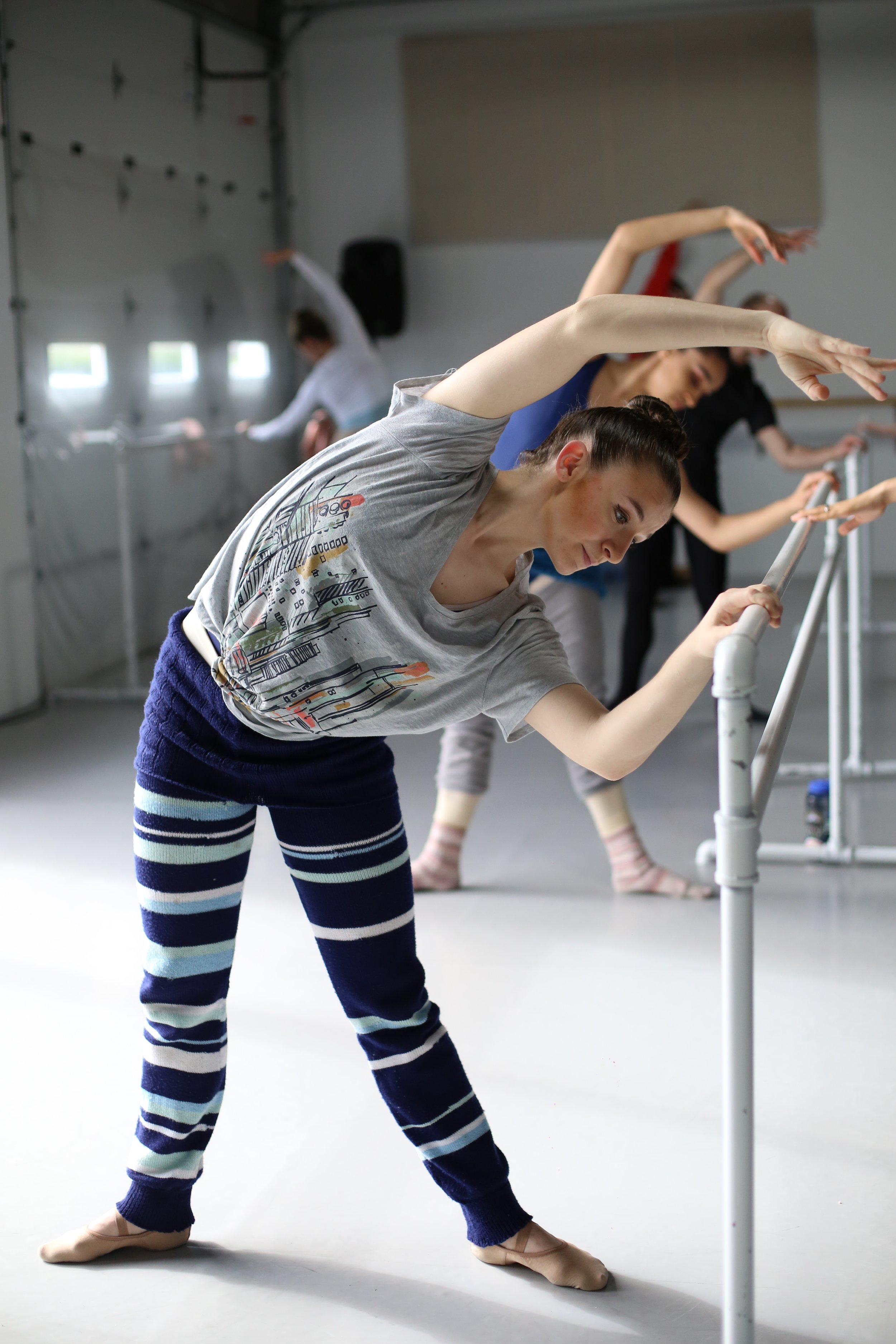Young woman practicing ballet at a barre in a dance studio, wearing striped leggings and a grey t-shirt.