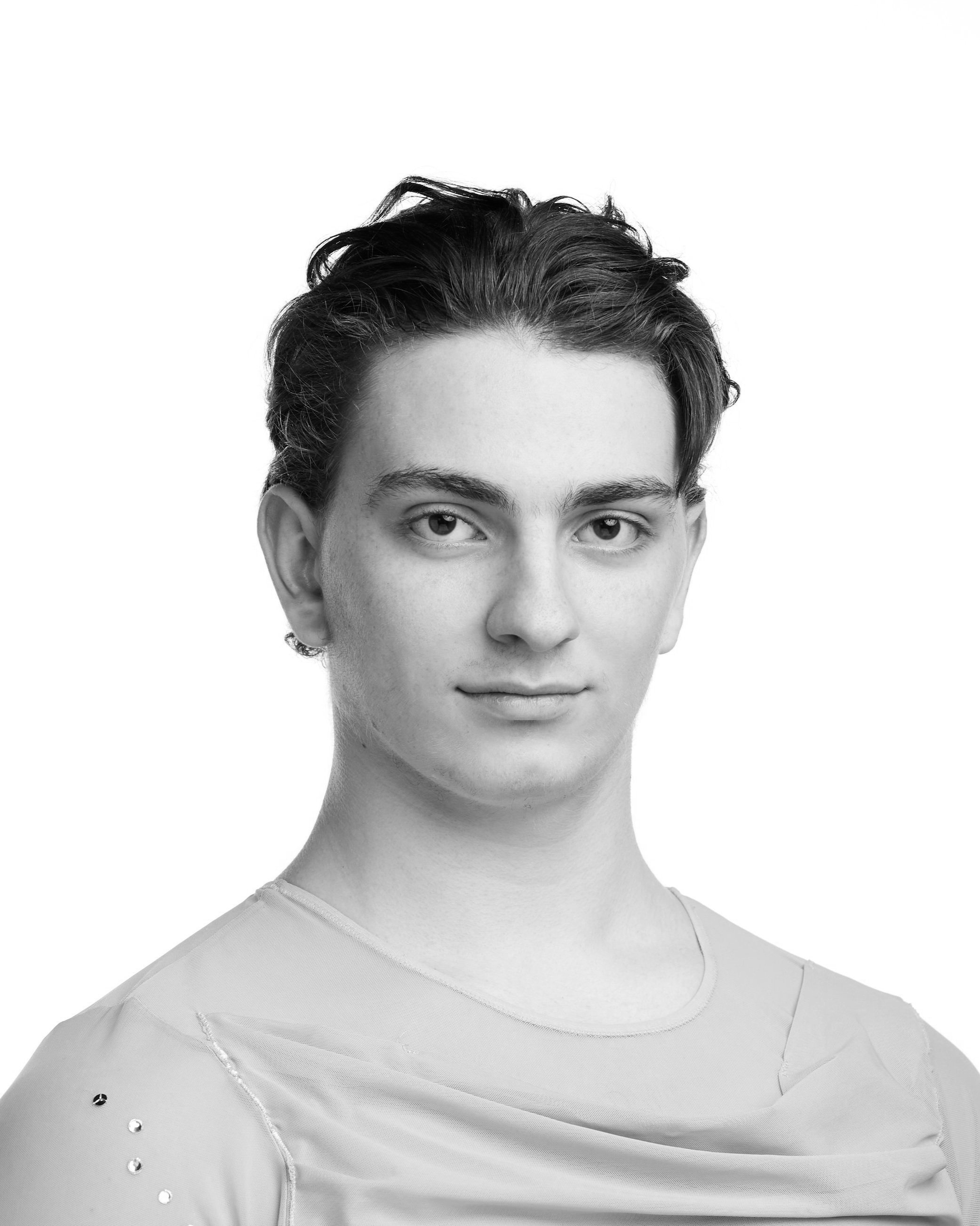 A black and white portrait of a young person with short dark hair, looking directly at the camera with a neutral expression, wearing a light-colored top with embellishments.