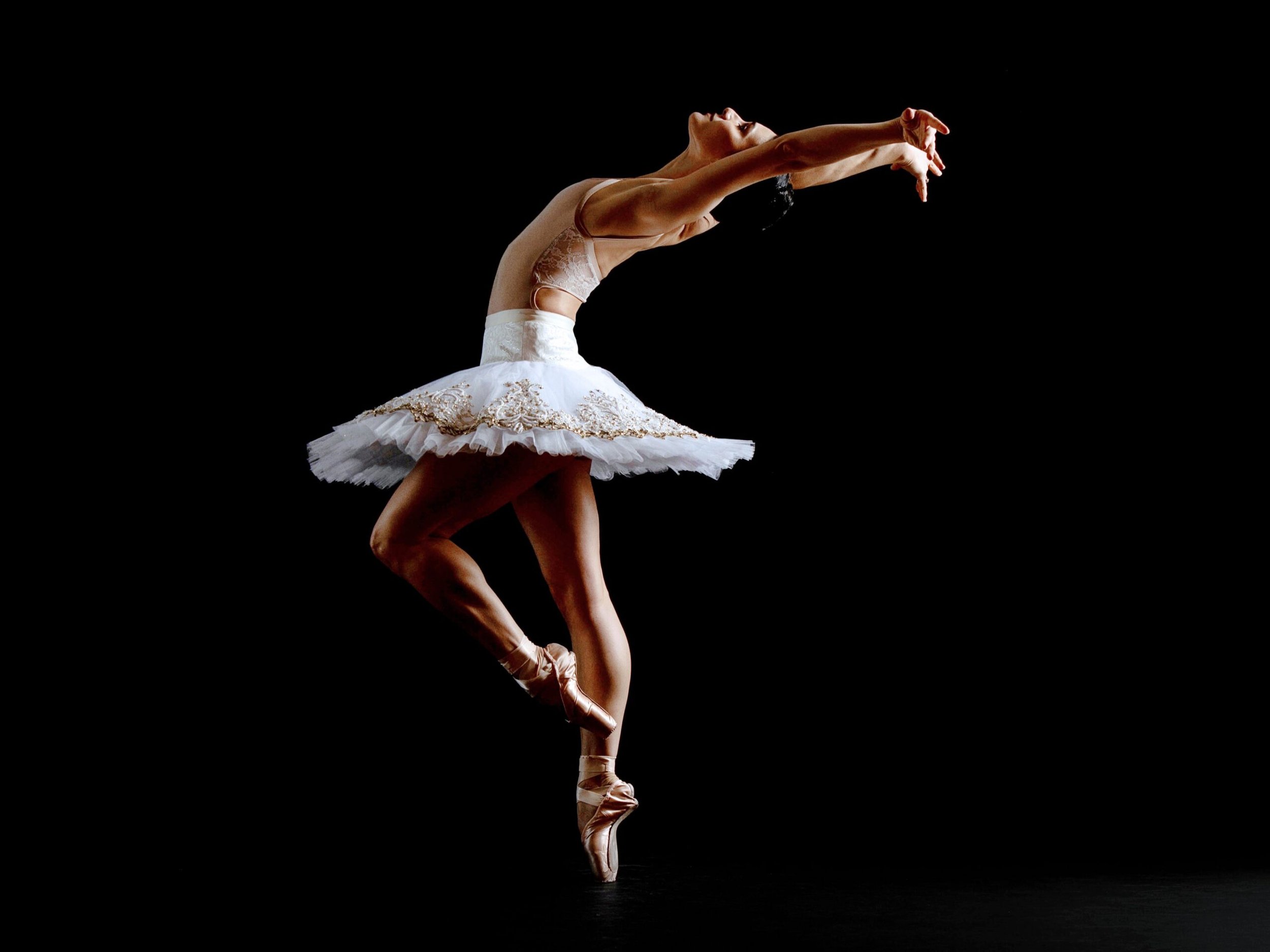A ballerina in a white tutu performing a dance pose against a black background.