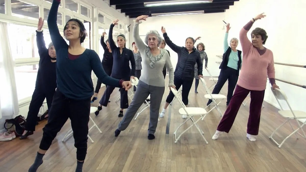 A group of women participating in a seated dance class, standing behind chairs and raising one arm in the air, in a bright studio with windows.